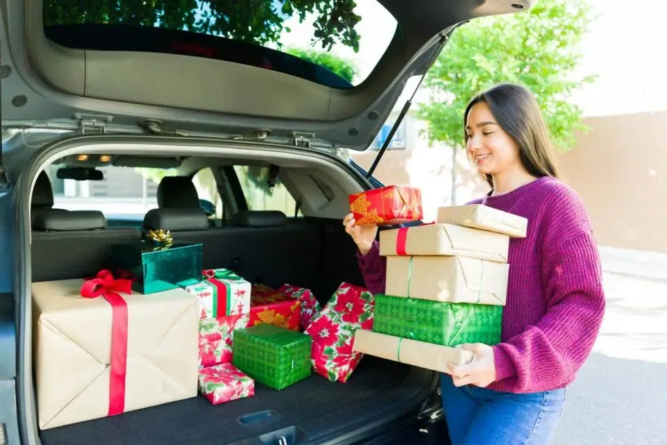 Happy beautiful woman carrying a lot of christmas presents from her car trunk after going shopping for the holidays