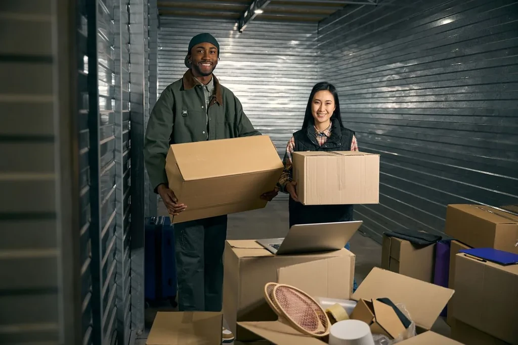Two people holding moving boxes inside a self storage unit aisle with stacked boxes and a laptop nearby.